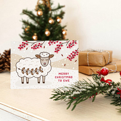 cute woolly sheep christmas card sat on a tabletop with a blurred christmas tree, foliage and gifts in the background.