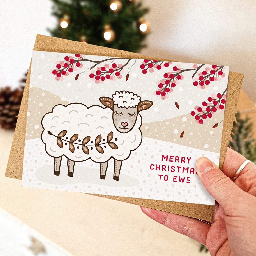 hand holding a cute sheep christmas card with kraft brown envelope with blurred christmas tree in the background.