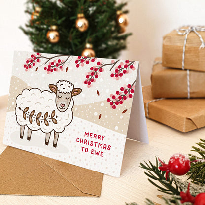 cute sheep christmas card with kraft brown envelope sat on a tabletop with gifts, foliage and a small christmas tree.