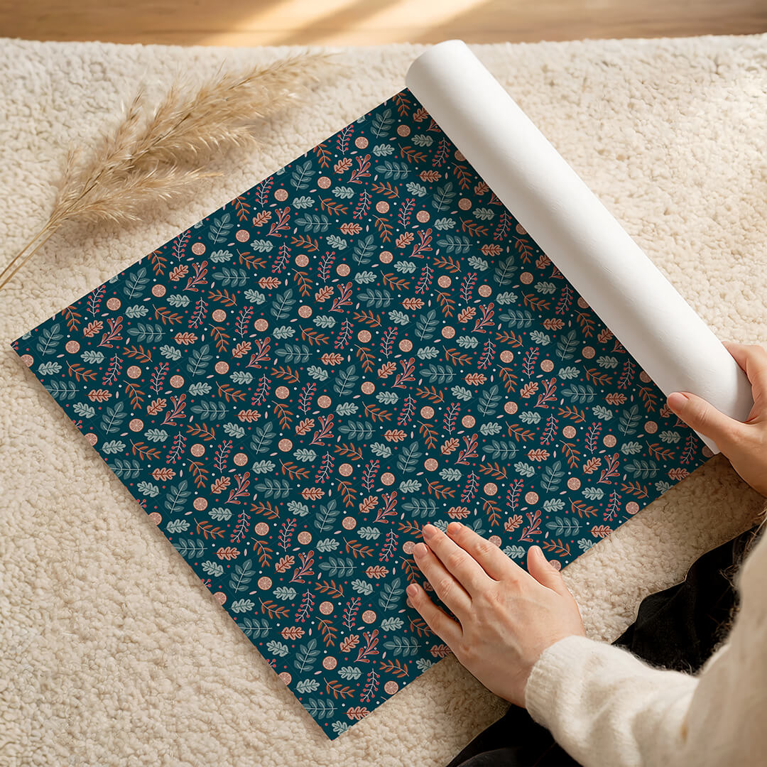 Person unrolling a roll of dark blue botanical Christmas wrapping paper on a fluffy cream rug.