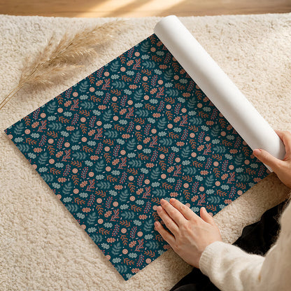 Person unrolling a roll of dark blue botanical Christmas wrapping paper on a fluffy cream rug.