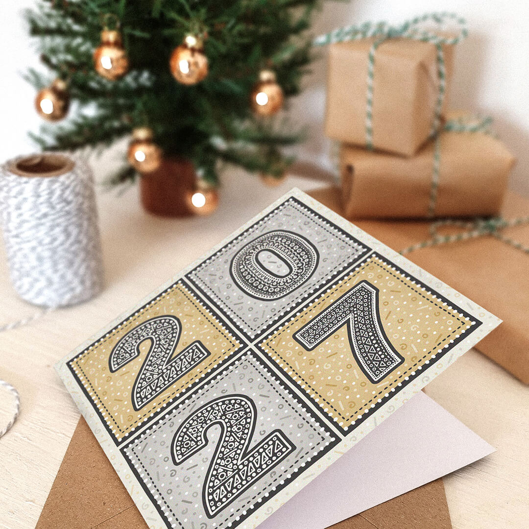 unique gold and silver 2027 new year card lying on white dresser with blurred christmas tree and gifts wrapped in kraft brown paper