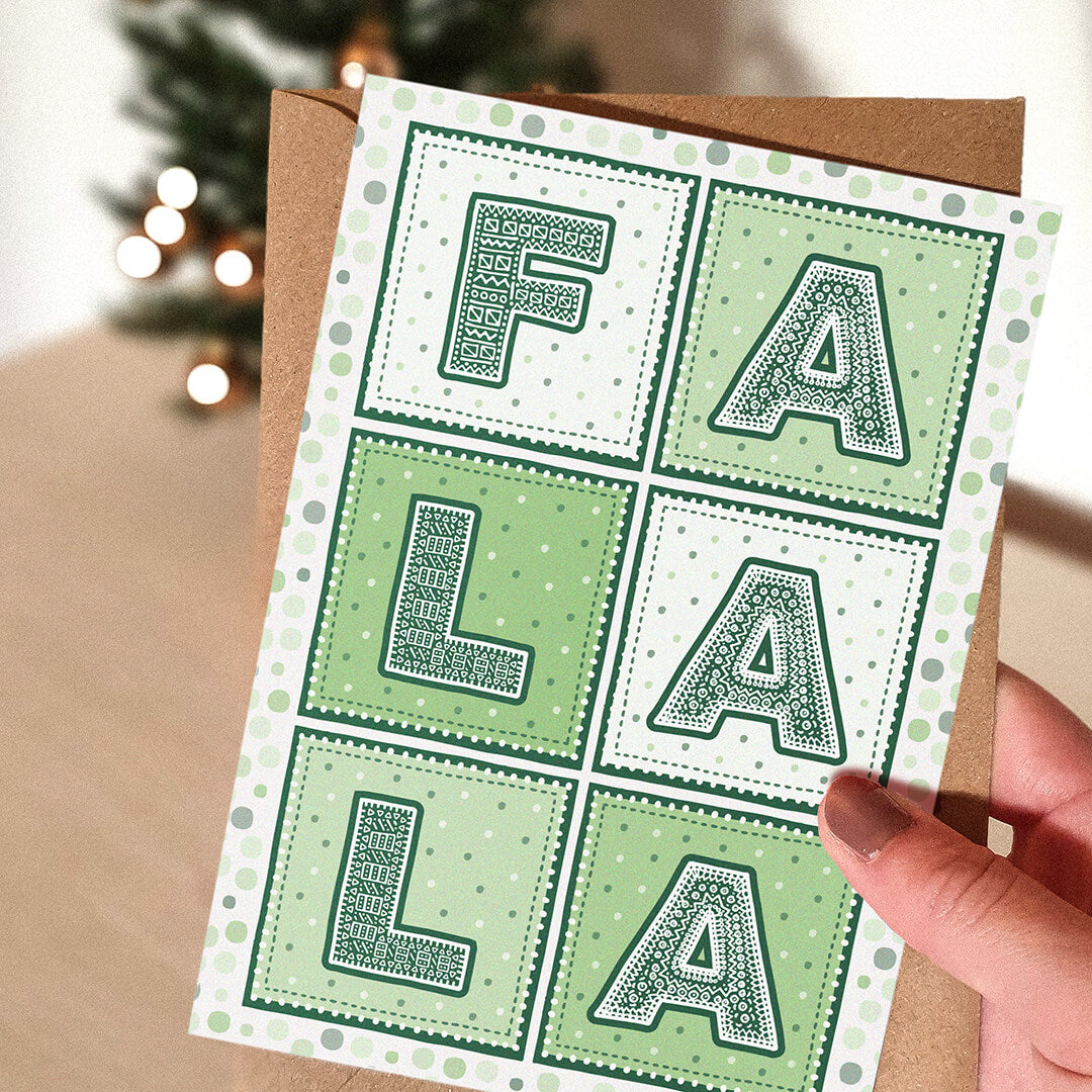 Green Christmas Card with patterned typography spelling 'FA LA LA' held by a hand with a Christmas tree in the background