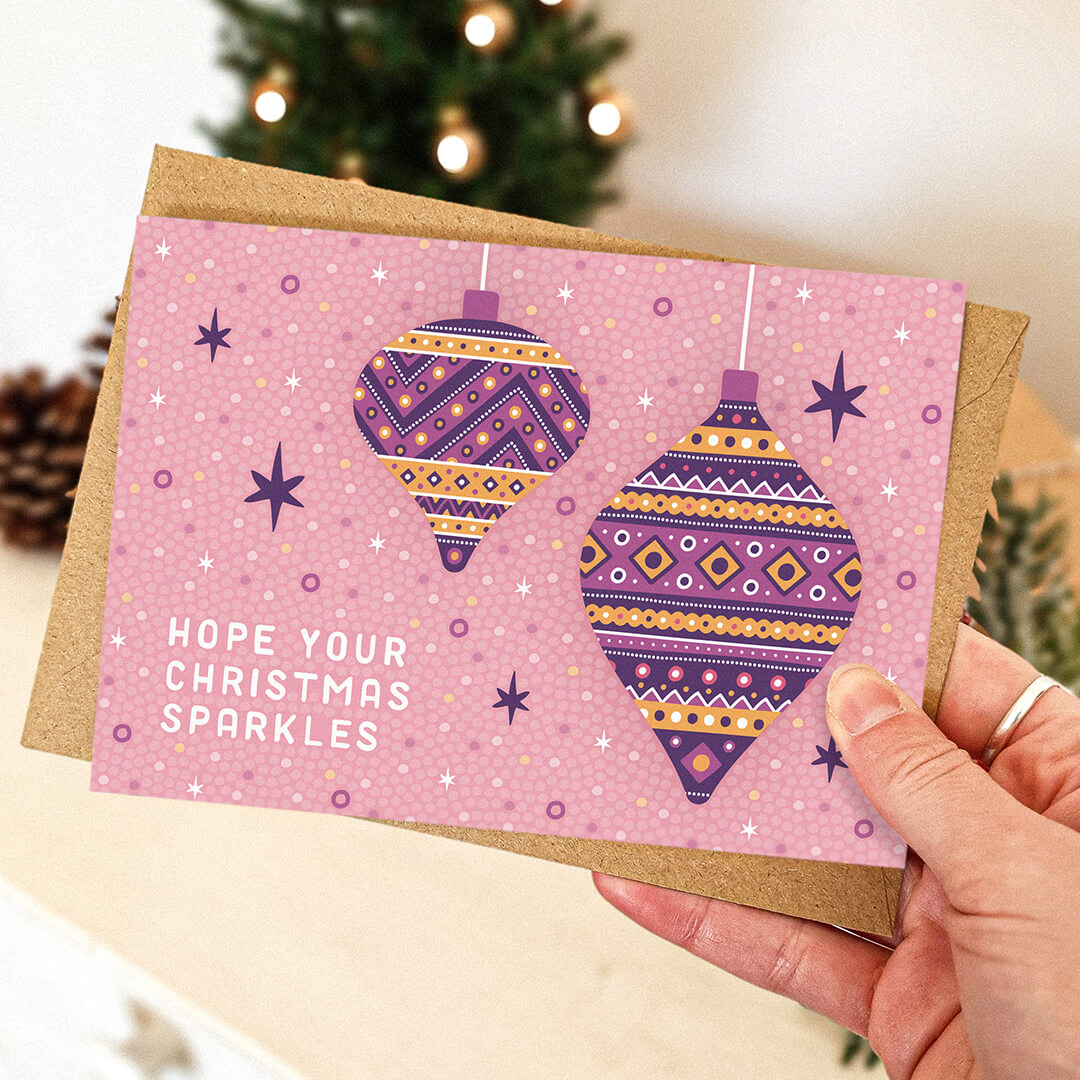 Hand holding light pink baubles Christmas card featuring gold and purple bauble illustrations and hope your Christmas sparkles message Blank inside Recycled kraft brown envelope