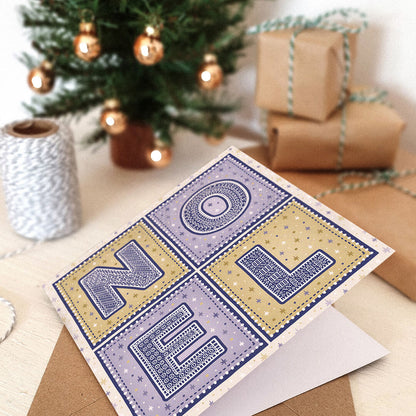 Purple and gold Noel Christmas card with 'NOEL' text in a festive setting with wrapped gifts and a small Christmas tree.