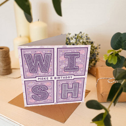 Purple birthday card with 'Make a Birthday Wish' text sat on a white wooden dresser with plants and candles in the background