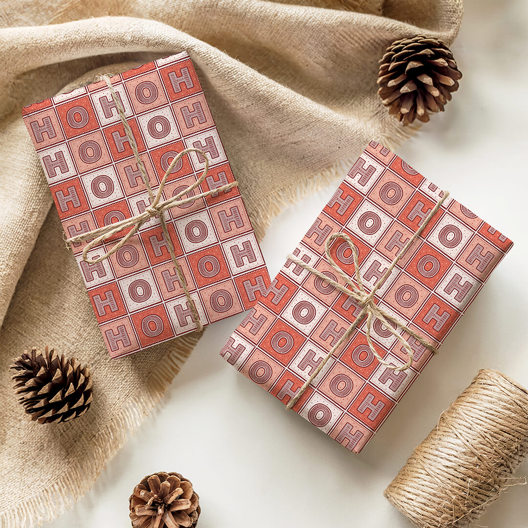 Two gift boxes with 'HO HO HO' Christmas gift wrap on a beige fabric background with pine cones and twine.