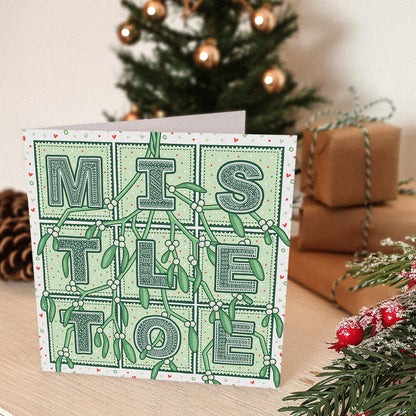 Romantic Mistletoe Christmas card with 'Mistletoe' typography and mistletoe illustrations in front of a decorated Christmas tree and presents.