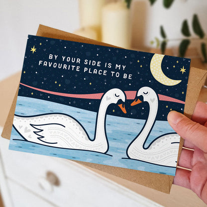 hand holding romantic anniversary card featuring two swans and a moonlit night sky and the message 'by your side is my favourite place to be' blank inside recycled kraft brown envelope