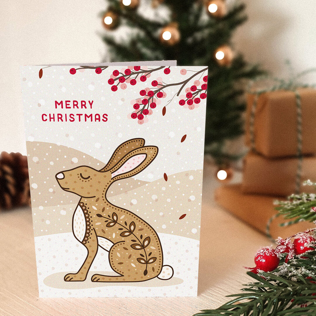 Christmas card with a rabbit or hare illustration and 'Merry Christmas' message, placed on a cream wooden table with festive decorations.