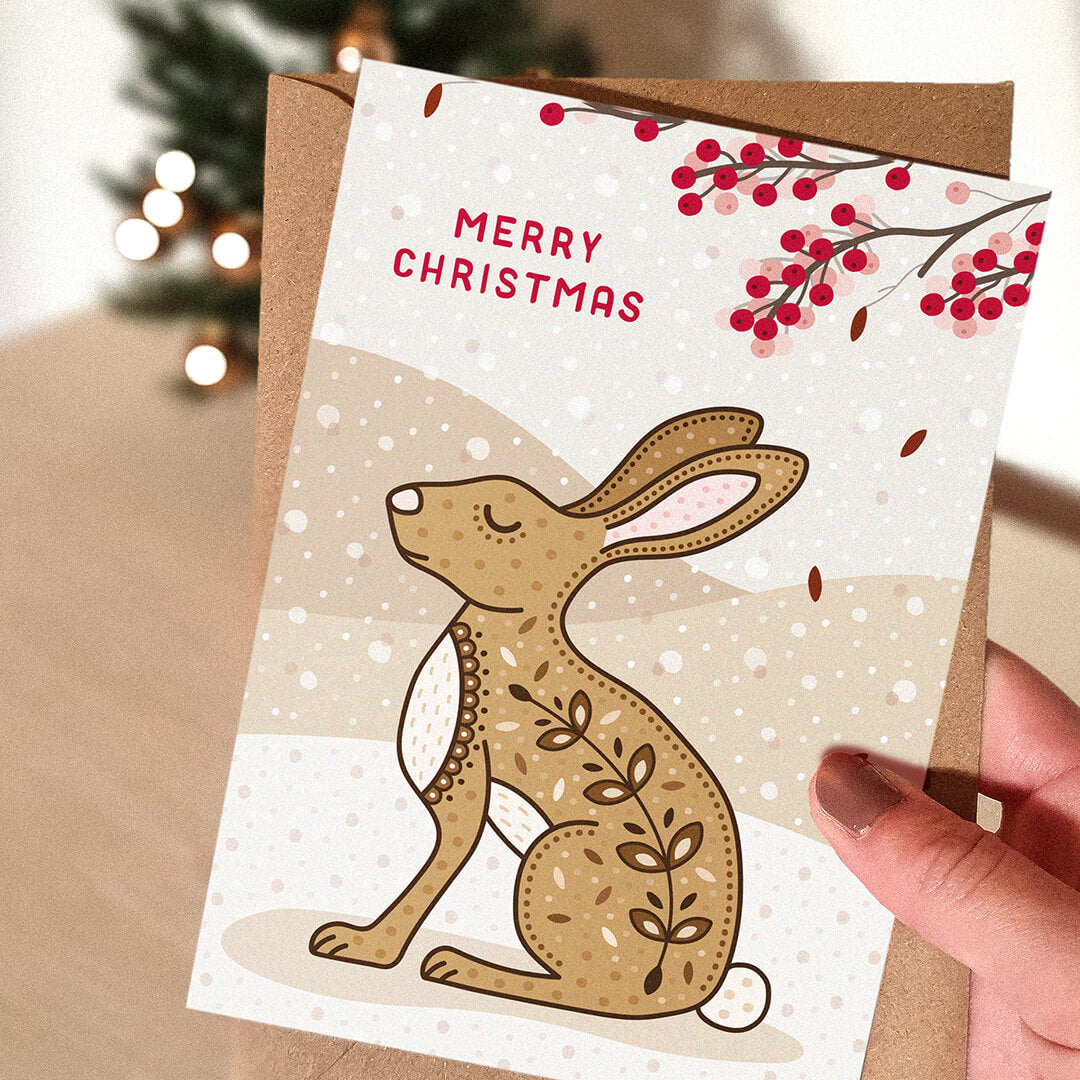 Hand holding a Christmas card with a rabbit or hare illustration and 'Merry Christmas' message.