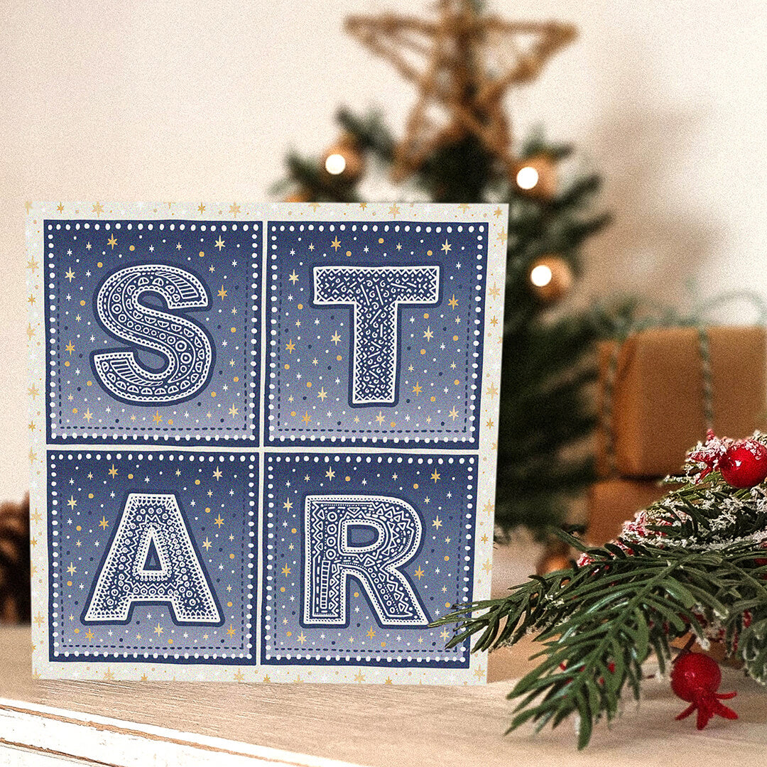 Star Christmas card with 'STAR' typography and star illustrations on a table with Christmas decorations in the background