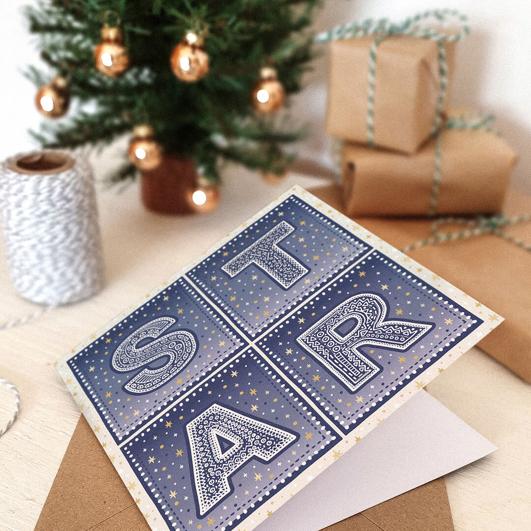 Star Christmas card with 'STAR' typography and star illustrations on a table with presents and a Christmas tree in the background