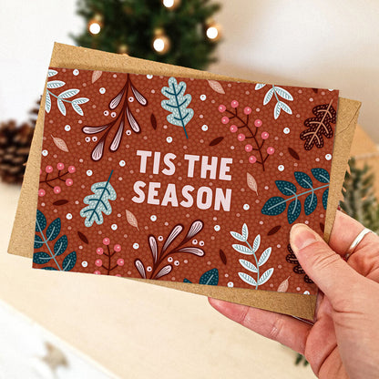 Hand holding unique tis the season red Christmas card with festive botanical illustrations of leaves, berries and foliage Printed on FSC certified card Supplied with kraft brown recycled envelope