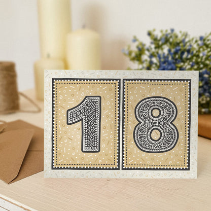 unique black and gold 18th birthday card sat on white wooden dresser with candles and flowers in the background