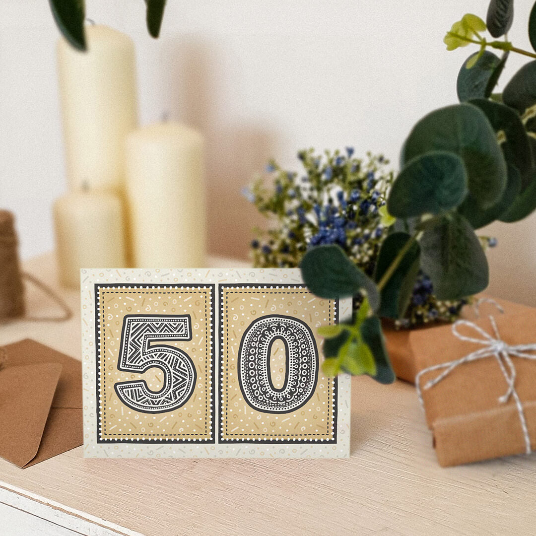 unique black and gold 50th birthday card sat on a white dresser with candles, flowers and gifts