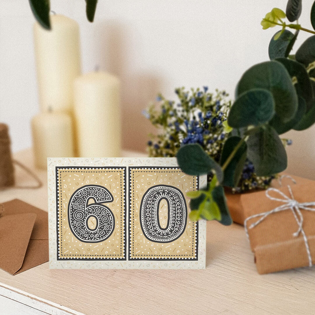 unique black and gold 60th birthday card sat on a wooden dresser surrounded by plants, gifts and candles