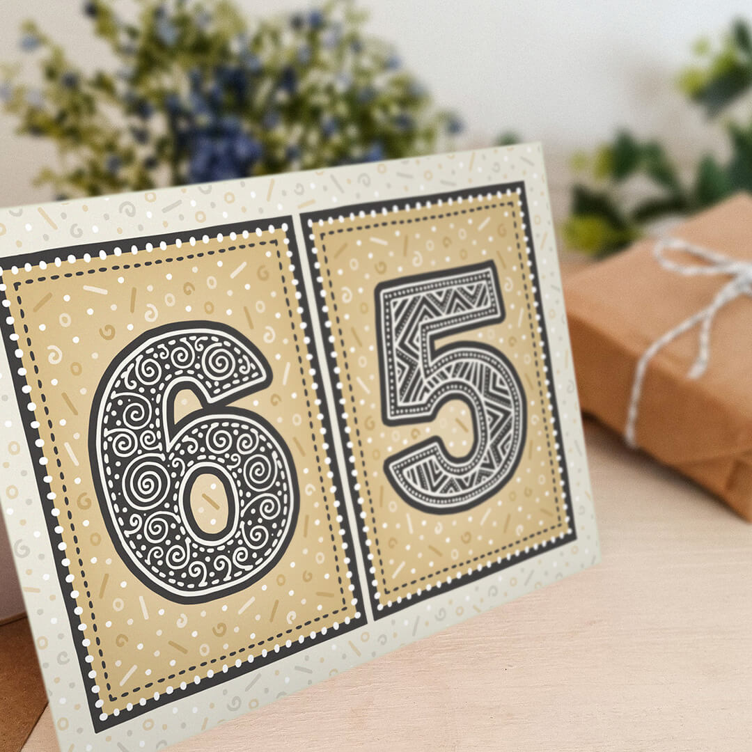 Gold 65th birthday card sat on a cream wooden shelf with blurred flowers and gifts in the background.