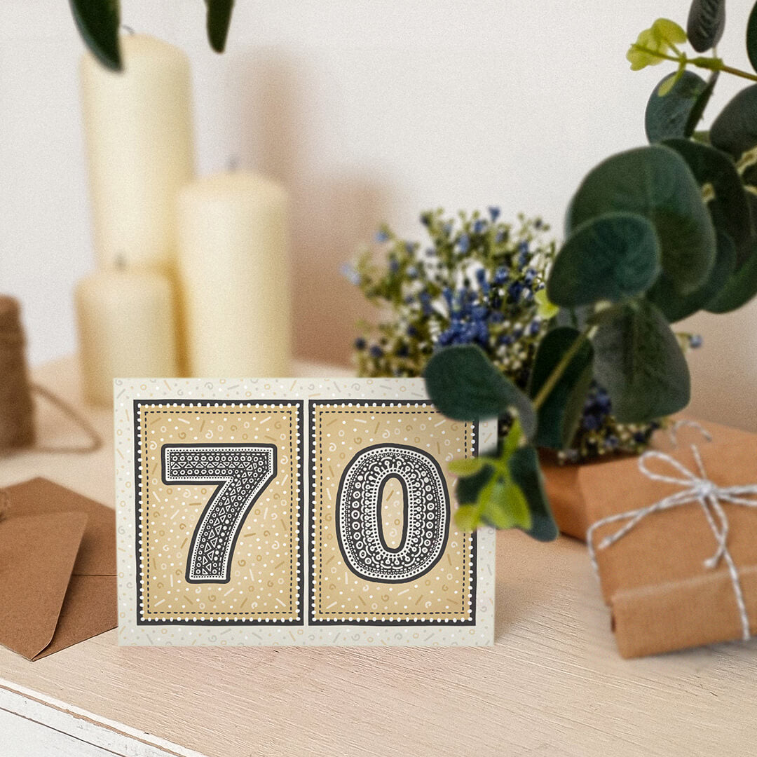 unique black and gold 70th birthday card sat on white dresser surrounded by plants, gifts and candles