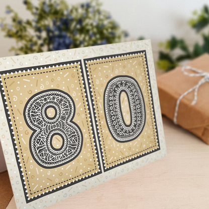unique black and gold 80th birthday card with flowers and gifts in the background