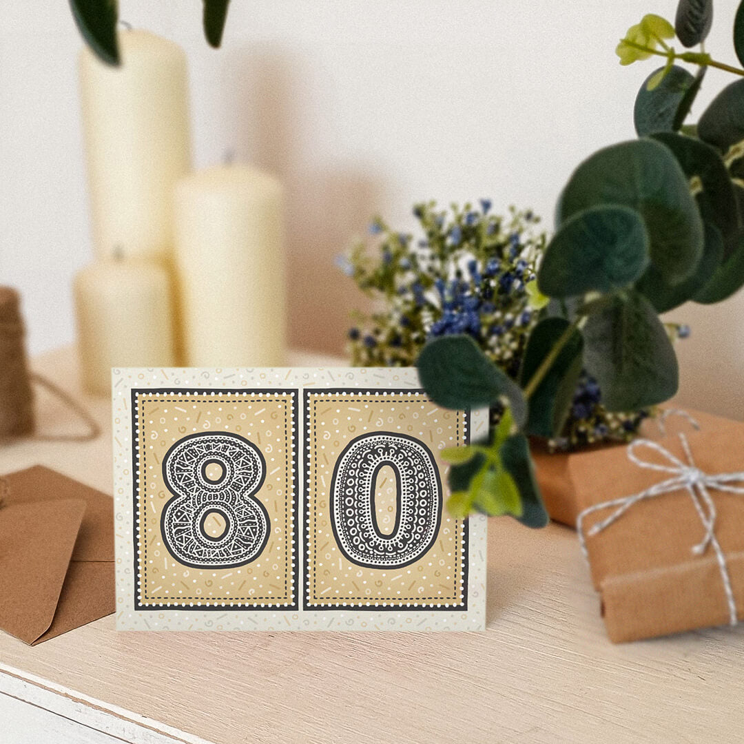 unique black and gold 80th birthday card sat on a white wooden dresser with candles, plants and gifts