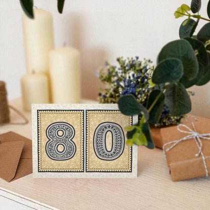 unique black and gold 80th birthday card sat on a white wooden dresser with candles, plants and gifts