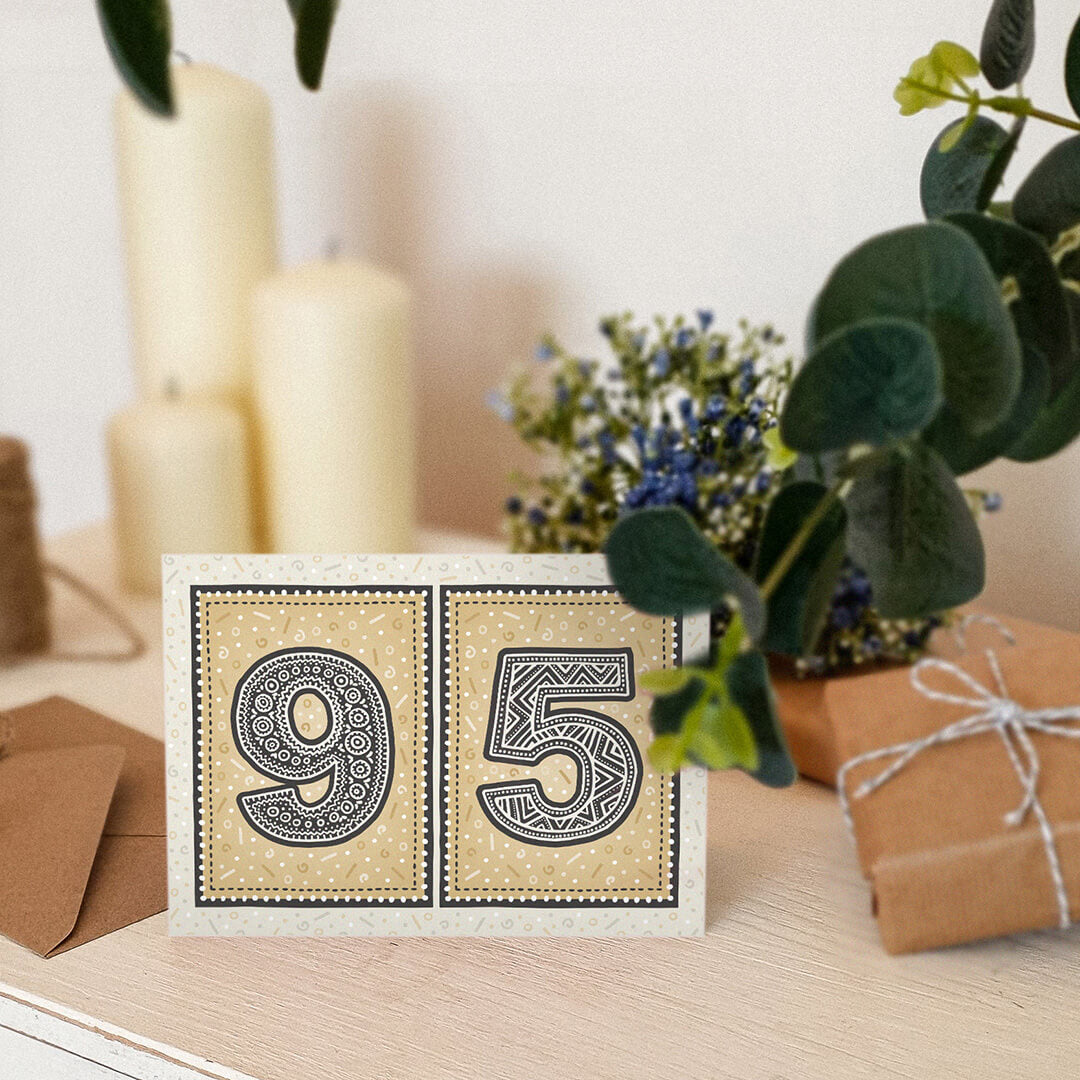 Gold 95th birthday card stood on a cream wooden surface with candles, plants and gifts.