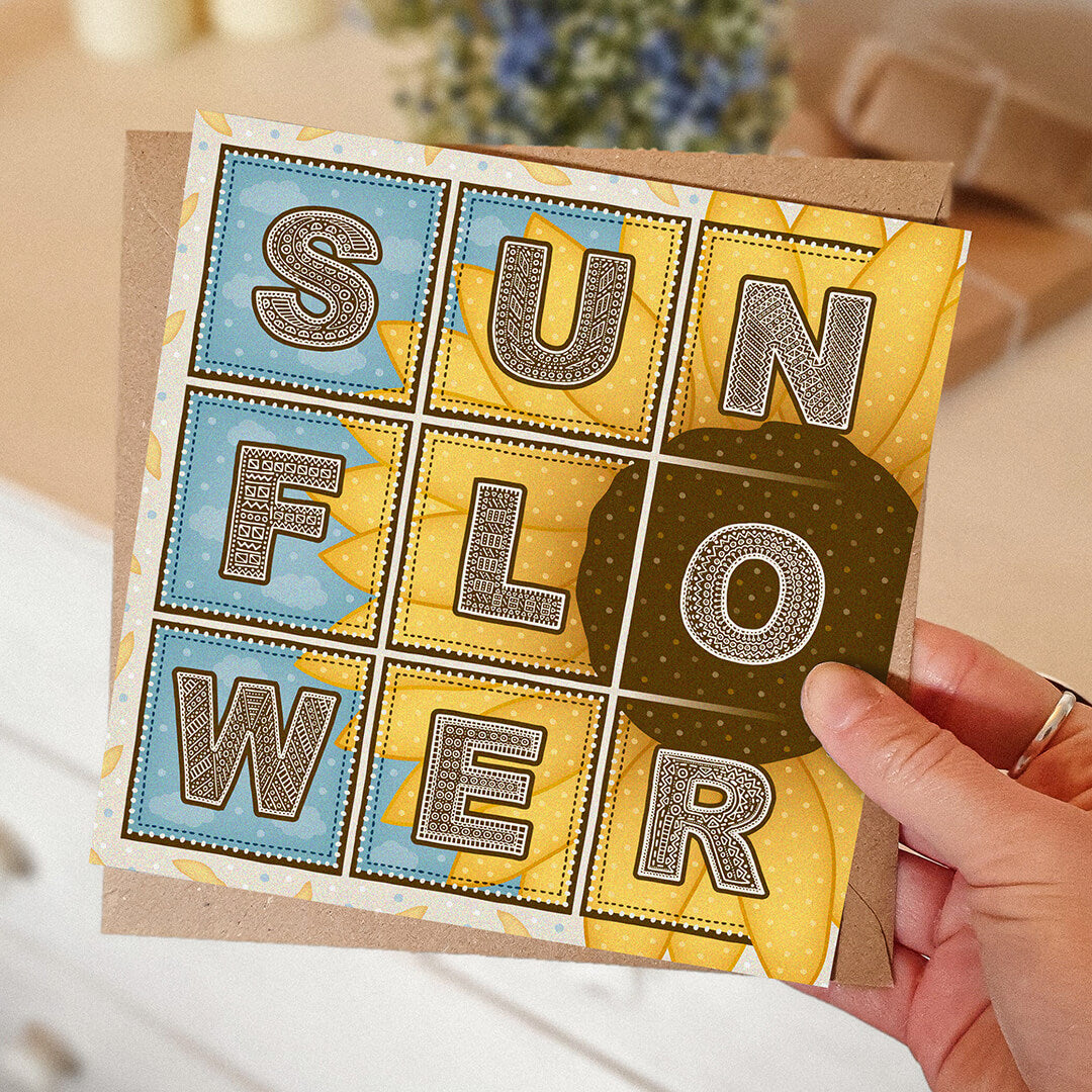 Hand holding a blue and yellow sunflower greeting card with 'Sunflower' text and sunflower illustration with a blurred background.