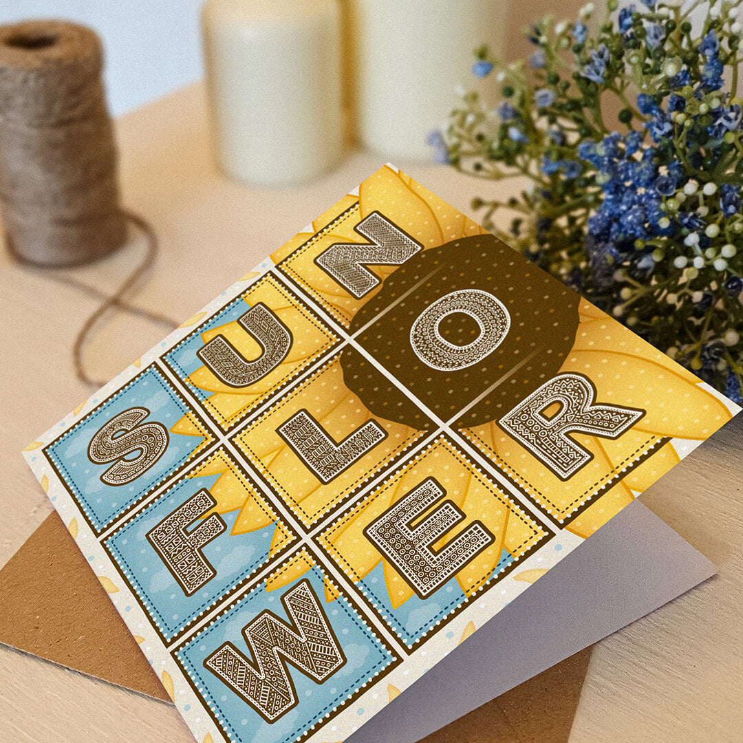 Blue and yellow sunflower greeting card with 'Sunflower' text and sunflower illustration on a white wooden dresser with blue flowers in the background.