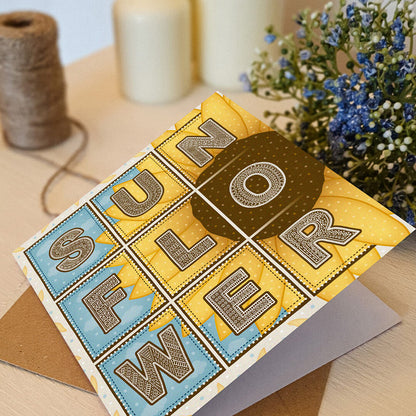 Blue and yellow sunflower greeting card with 'Sunflower' text and sunflower illustration on a white wooden dresser with blue flowers in the background.