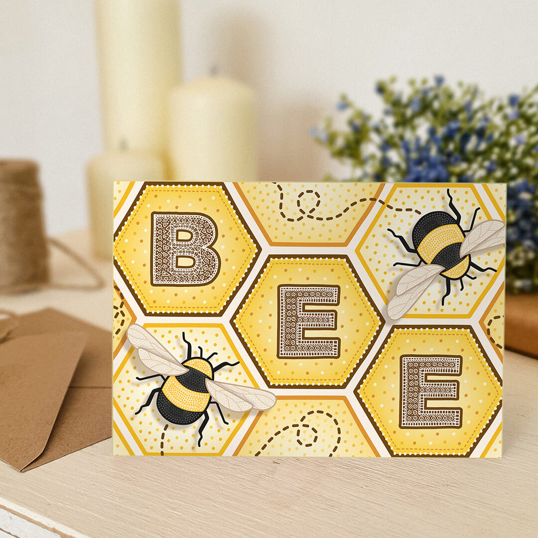 Bee greeting card with bee illustrations, detailed typography and honeycomb design on a white wooden dresser with plants and candles in the background