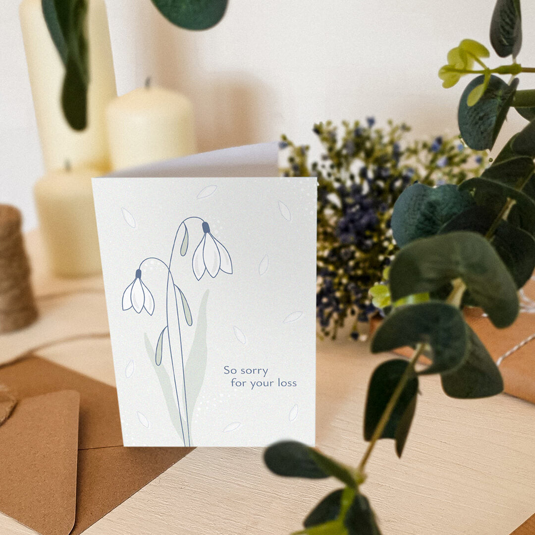 white snowdrops sympathy card sat on a white dresser surrounded by plants and candles