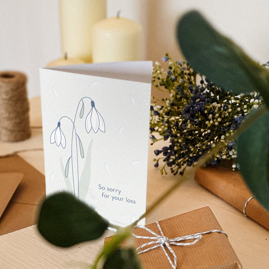 white snowdrops sympathy card sat on a white dresser surrounded by flowers, candles and gifts